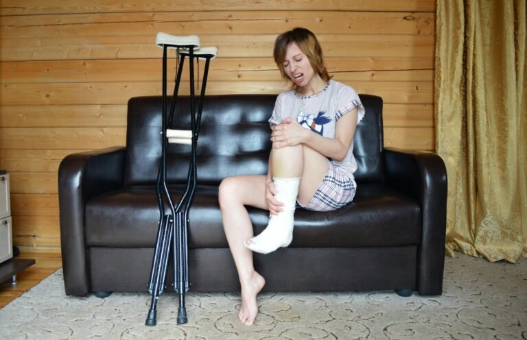 Full body of woman with aching leg sitting on couch near pair of crutches