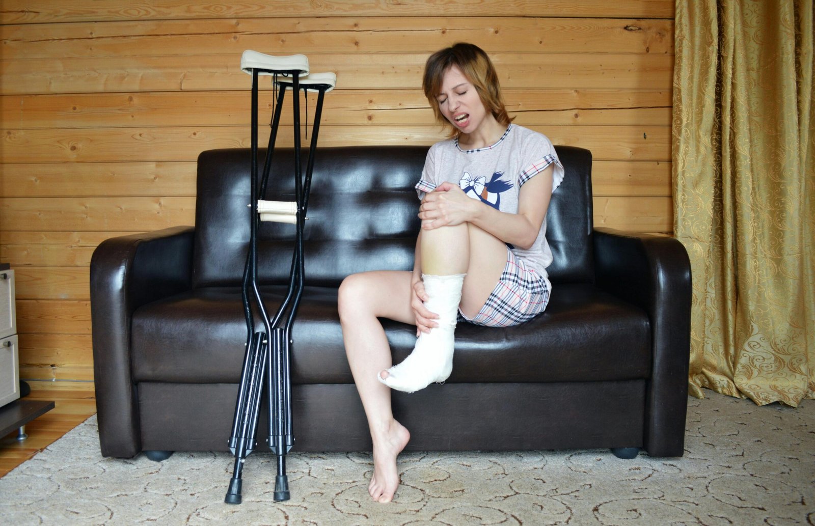 Full body of woman with aching leg sitting on couch near pair of crutches
